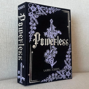 Könnte beinhalten: Ein Hardcover-Buch mit dem Titel "Powerless" von Lauren Roberts. Das Buch hat einen schwarzen Einband mit einem floralen und Schwert-Design in Weiß. Der Buchrücken zeigt ebenfalls den Titel und den Namen des Autors.