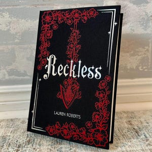 Könnte beinhalten: Ein Hardcover-Buch mit dem Titel "Reckless" von Lauren Roberts. Das Buchcover ist schwarz mit einem roten Blumendesign und silbernen Akzenten. Der Titel ist in einer dekorativen weißen Schrift.