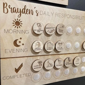 May include: A wooden chart with the text "Brayden's Daily Responsibilities" and a checklist of daily tasks for children. The chart has a sun icon for morning, a moon icon for evening, and a checkmark for completed tasks. The tasks include getting dressed, brushing teeth, unpacking a backpack, cleaning up blankets, putting up laundry, cleaning the bedroom, vacuuming, showering, cleaning the bathroom, and doing dishes.
