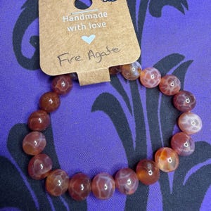May include: A beaded bracelet made of round, translucent fire agate stones in shades of orange, red, and brown. The bracelet is attached to a rectangular brown cardboard display tag with the text Handmade with love, a heart icon, and Fire Agate.