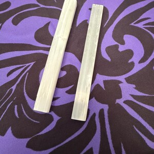 Peut inclure: Deux baguettes rectangulaires en cristal de sélénite blanc cassé sont présentées sur un fond violet avec un motif floral marron foncé. Les baguettes sont positionnées en diagonale, mettant en valeur leur texture lisse et translucide.