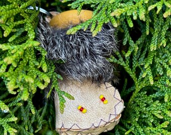 Puede incluir: Un pequeño adorno de mocasín hecho a mano al estilo nativo americano. El mocasín está hecho de cuero marrón con un ribete de piel y está decorado con cuentas rojas y amarillas. Cuelga entre ramas verdes de hoja perenne.