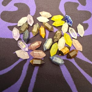 Puede incluir: Una colección de pequeños cristales de doble terminación en varios colores, incluyendo transparente, amarillo, rosa, marrón y azul. Los cristales están dispersos sobre una superficie con un patrón morado y negro, creando una disposición visualmente atractiva.