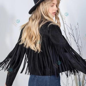 Puede incluir: Una chaqueta negra con largos flecos a lo largo de las mangas y la espalda, combinada con un sombrero fedora negro y pantalones vaqueros azules. La chaqueta tiene una textura suave y largos flecos fluidos que salen de los hombros y brazos.