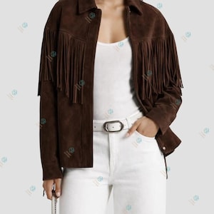 Peut inclure: Une veste en daim marron foncé avec de longues franges sur la poitrine et les manches, portée avec un débardeur blanc, un pantalon blanc et une ceinture blanche à boucle ovale argentée. La veste présente un col classique et des manches longues.