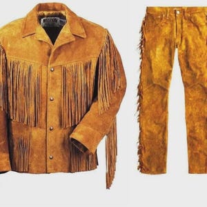 May include: A tan suede jacket and matching pants set. The jacket features a pointed collar, four metal snap buttons, and long fringe detailing across the chest, back, and sleeves. The pants include matching fringe along the outer leg seams.