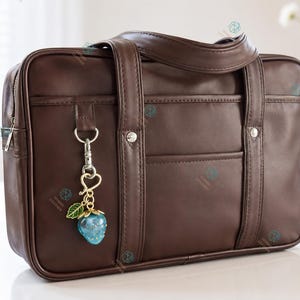 Può includere: Una borsa rettangolare in pelle marrone scuro con due manici superiori, uno scomparto principale con cerniera e una tasca frontale. Un portachiavi argentato con una foglia verde e un ciondolo a forma di fragola blu pende da una cinghia.