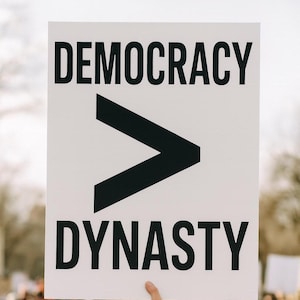 以下が含まれることがあります： 「DEMOCRACY」と「DYNASTY」という言葉が太字の黒い文字で書かれた白い看板。大きな黒い大なり記号が単語の間にあります。看板は人が持っており、背景はぼやけています。