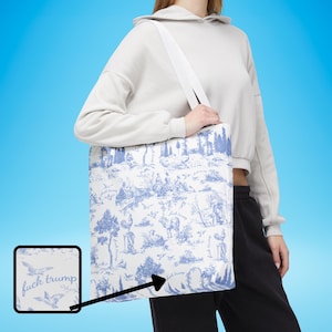 Puede incluir: Bolso tote blanco con un estampado de toile de Jouy azul que representa figuras, árboles y animales. El bolso tiene una correa blanca y las palabras "fuck trump" están impresas en el bolso.