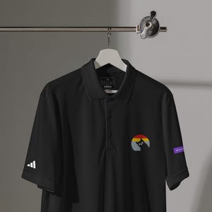 Puede incluir: Polo negro con cuello y mangas cortas. La camisa presenta un pequeño diseño bordado de una silueta de gato dentro de un círculo arcoíris en el pecho. El logo de Adidas está en la manga izquierda.