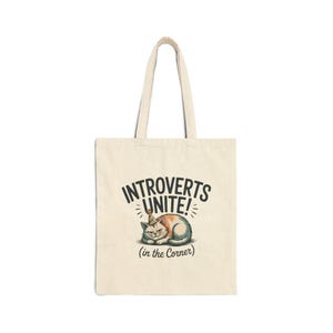 May include: A cream-coloured tote bag featuring the text "INTROVERTS UNITE!" and a cartoon cat wearing a party hat. The cat is orange, white, and brown, with the text "(in the Corner)" below. The bag has two handles.