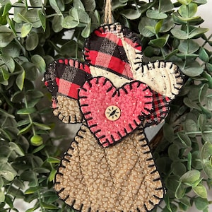 May include: A handmade angel ornament with a heart-shaped body and wings. The wings are made of red and black plaid fabric, and the body is a pink heart with a wooden button. The angel is stitched with black thread and has a jute string for hanging.
