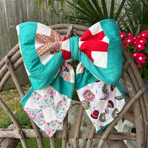 Puede incluir: Un gran lazo hecho a mano con un edredón de retazos. El lazo presenta una mezcla de patrones y colores, incluyendo cuadrados rojos y blancos, estampados florales y bordes verde azulado. Se exhibe en una silla de madera rústica.