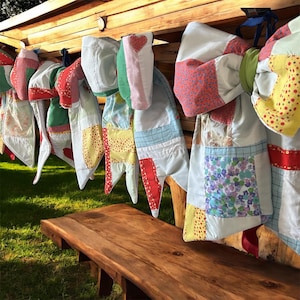 May include: A collection of colorful, quilted oven mitts hanging on a wooden structure. The mitts feature various patterns, including polka dots, floral prints, and hearts, with red, yellow, and blue accents. A wooden bench sits below.