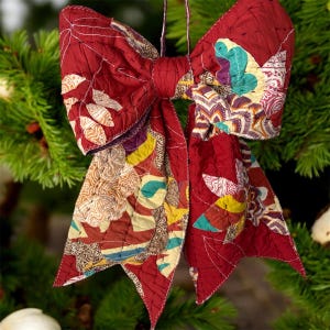 May include: A decorative bow ornament crafted from quilted fabric in a deep red hue. The surface features a patchwork of intricate patterns in shades of yellow, teal, purple, and white, with visible stitching throughout the entire textile construction.