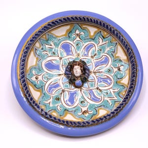 May include: A round, ceramic decorative plate with a light blue border and a detailed floral design in shades of blue, teal, and yellow. The center features a sculpted face with a blue gem. The plate has a diameter of approximately 10 inches.