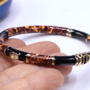 May include: A bamboo-style bracelet with a tortoiseshell pattern, black accents, and gold-tone metal details. The bracelet features small, clear gemstones and is a stylish accessory.