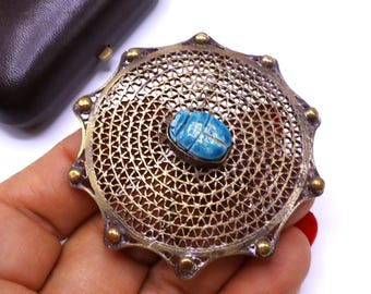 May include: Vintage round brooch with a filigree design. The brooch features a central, light blue scarab beetle. The outer edge has small gold-colored accents. The brooch is made of a gold-colored metal.