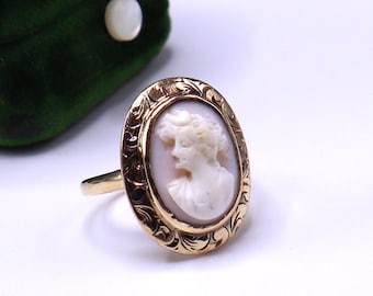 May include: A gold-toned ring featuring an oval cameo with a profile of a woman. The cameo is set in a decorative, engraved frame. The ring's band is a simple, polished gold. The cameo has a light pink background and a white relief.