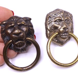 May include: Two metal lion head door knockers with circular rings. One lion head is a dark bronze tone, and the other is a lighter, weathered brass tone. Both feature detailed manes and facial features with a ring held in the lion's mouth.