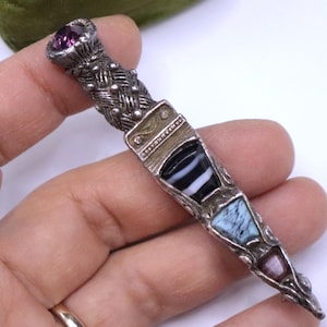 May include: A silver-toned metal brooch shaped like a dagger, featuring a textured handle topped with a round purple stone. The blade is inlaid with three distinct stones: a black and white striped stone, a light blue marbled stone, and a small purple stone.
