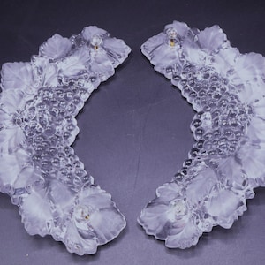 May include: Two clear glass grape-shaped decorative pieces. Each piece features a cluster of round, textured grapes with surrounding leaves. The glass has a frosted appearance, and the pieces are set against a dark background.