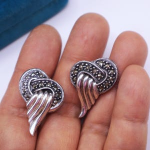 May include: A pair of vintage silver-toned earrings. Each earring features a heart-shaped design with marcasite stones and a stylized wing detail. The earrings are displayed on a light background.