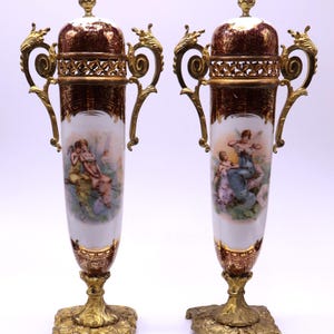 May include: A pair of tall porcelain urns with lids, featuring deep burgundy and gold decorative bands, ornate gold-toned metal handles, and square bases. Each urn displays a central hand-painted scene of figures in a garden setting.