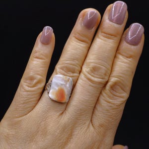 May include: A silver ring with a rectangular, light-colored stone featuring orange and white patterns. The ring is on a hand with painted nails. The ring's setting has decorative details.