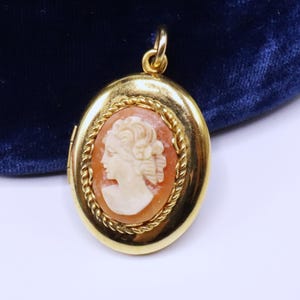 May include: Oval gold-tone locket with a cameo of a woman's profile. The cameo is set in a reddish-orange background and framed with a twisted rope design. The locket has a loop at the top for a chain.
