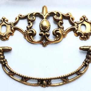 May include: A gold-toned metal drawer pull consisting of two separate pieces: a curved, ornate handle with textured detailing and a matching decorative backplate featuring intricate scrollwork and two circular mounting holes.