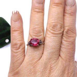 May include: A silver ring featuring a large, oval-cut, bright red gemstone. The ring has an ornate, filigree-style setting. The ring is shown on a hand with manicured nails.