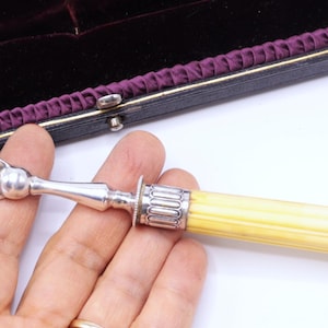 May include: An antique silver serving fork with a decorative handle. The fork has three tines and ornate detailing on the head. The handle is a light yellow color. The fork is displayed in a black case with a purple interior.
