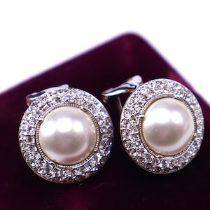 May include: A pair of round cufflinks with a central pearl surrounded by a ring of small, clear crystals. The cufflinks have a silver-colored metal backing and are displayed on a dark purple velvet surface.