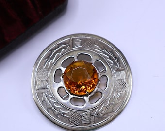 May include: A silver-toned brooch with a large, faceted amber-colored gemstone at its center. The brooch features a circular design with engraved thistle motifs around the edge, showcasing a vintage or antique style.
