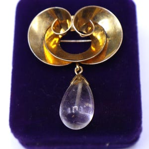 May include: A gold-toned brooch with a swirling design and a clear, teardrop-shaped pendant. The brooch has a pin closure and is displayed on a purple velvet surface. The pendant is transparent and reflects light.