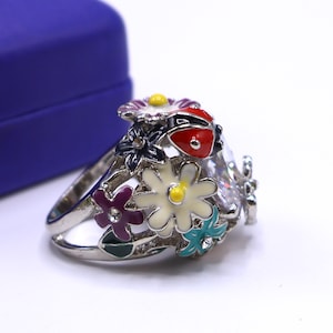 May include: A silver ring featuring a floral design with colorful enamel flowers and a red ladybug. The ring has a large, clear gemstone and is set against a white background.