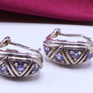 May include: A pair of silver-toned hoop earrings. Each earring features a geometric design with triangular patterns and small, round, purple gemstones. The earrings are displayed against a white background with a burgundy fabric in the background.