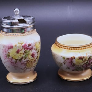 May include: A vintage cream-colored ceramic sugar bowl with a silver lid and a matching open bowl. Both pieces feature floral designs in pink, yellow, and green, with gold trim. The sugar bowl has a small spoon slot.