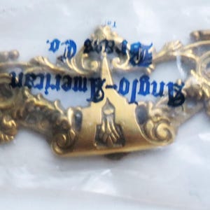 May include: A gold-toned metal drawer pull plate with ornate scrollwork and decorative detailing. The piece is enclosed in a clear plastic bag featuring the blue printed text Anglo-American Brass Co. The hardware has two circular mounting holes.