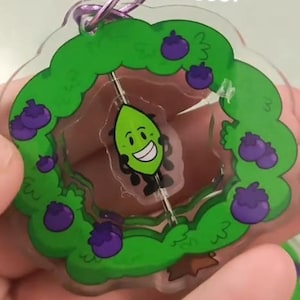 Puede incluir: Un llavero acrílico transparente con un personaje verde con forma de hoja sonriente, rodeado de un arbusto verde con bayas moradas. El llavero tiene un anillo plateado y el texto "if I sold these?"