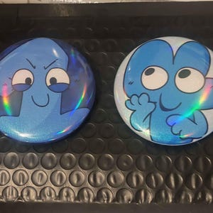 May include: Two round, blue character buttons with holographic glitter effects. One button features a blue number 1 with an angry face, and the other has a blue number 4 with a friendly expression. The buttons are on a black bubble mailer.