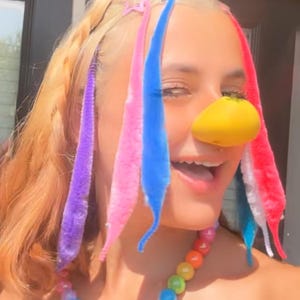 May include: A person with blonde hair styled with colorful pipe cleaners and star-shaped hair clips. A yellow nose and a rainbow beaded necklace complete the playful look. The image captures a fun, whimsical aesthetic.