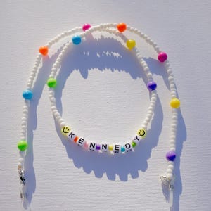 May include: White beaded eyeglass chain with colorful beads and the name "KENNEDY" spelled out in letter beads. The chain has a smiley face bead on each end of the name and is designed to hold eyeglasses.