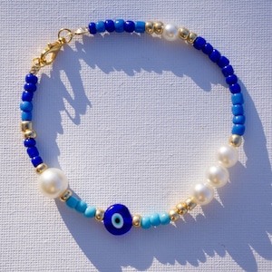 May include: A beaded bracelet featuring a blue evil eye charm, white faux pearls, and gold-colored beads. The bracelet has a gold-colored clasp and is composed of various shades of blue beads.