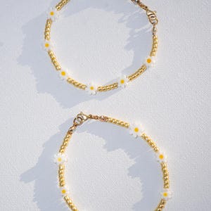 Op de afbeelding: Twee madeliefjeskettingarmbanden. Elke armband heeft witte madeliefjeskralen afgewisseld met goudkleurige kralen. De armbanden hebben goudkleurige sluitingen en worden op een wit oppervlak getoond.