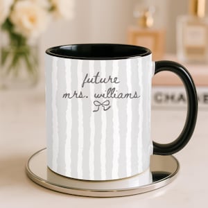 May include: A white ceramic mug with a black handle and interior, featuring a gray and white striped pattern. The mug has the text "future mrs. williams" in gray script with a bow graphic. The mug sits on a silver-colored coaster.