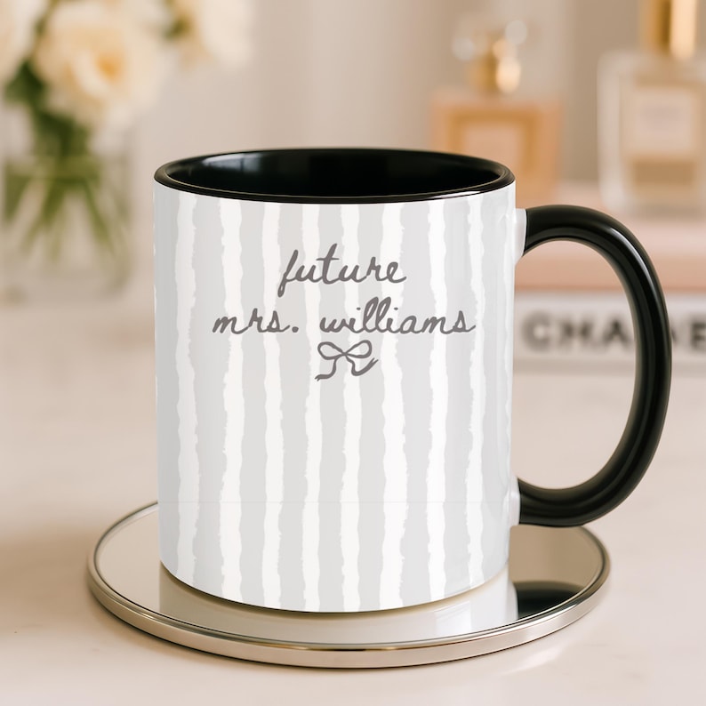 May include: A white ceramic mug with a black handle and interior, featuring a gray and white striped pattern. The mug has the text "future mrs. williams" in gray script with a bow graphic. The mug sits on a silver-colored coaster.