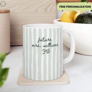 May include: White ceramic mug with a green and white striped pattern. The mug features the text "future mrs. williams" in a handwritten style, with a bow graphic. The mug is on a beige coaster.