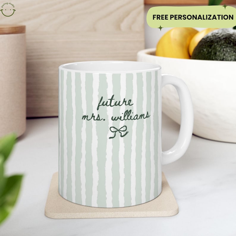 May include: White ceramic mug with a green and white striped pattern. The mug features the text "future mrs. williams" in a handwritten style, with a bow graphic. The mug is on a beige coaster.